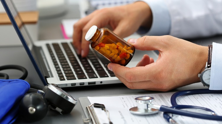Doctor holds and reads bottle of medication while sitting at his computer